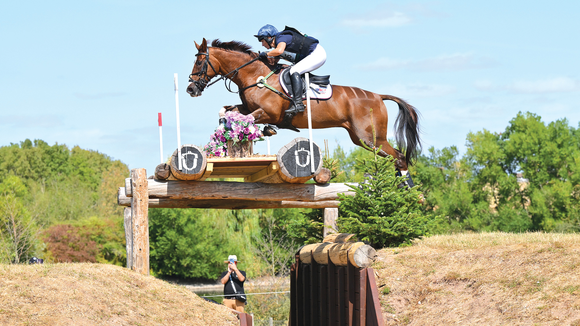 A chestnut horse jumps over a large oxer with a filled in top over a deep ditch at Hartpury. The loga have the Hartpury acorn logo on their ends.