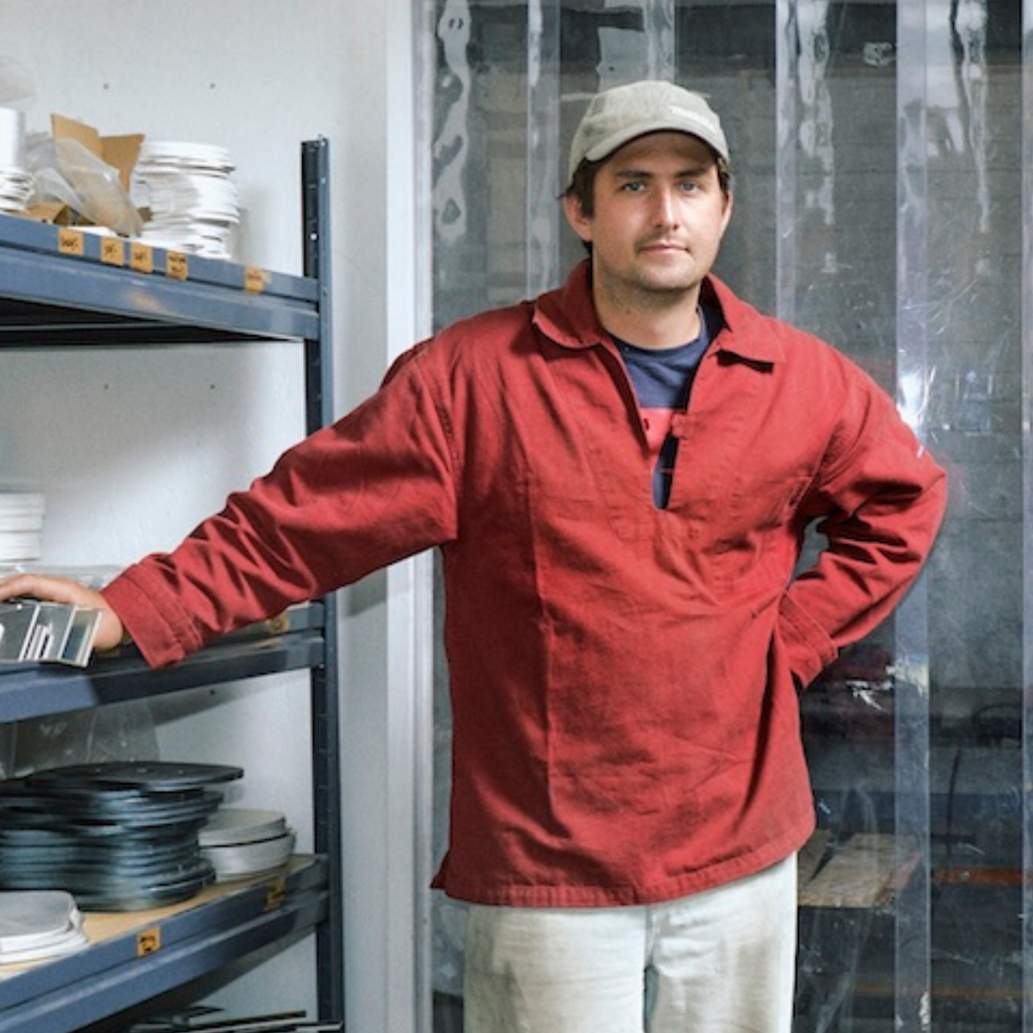 A man leaning on a storage rack in a red shirt, white pants, and a cap