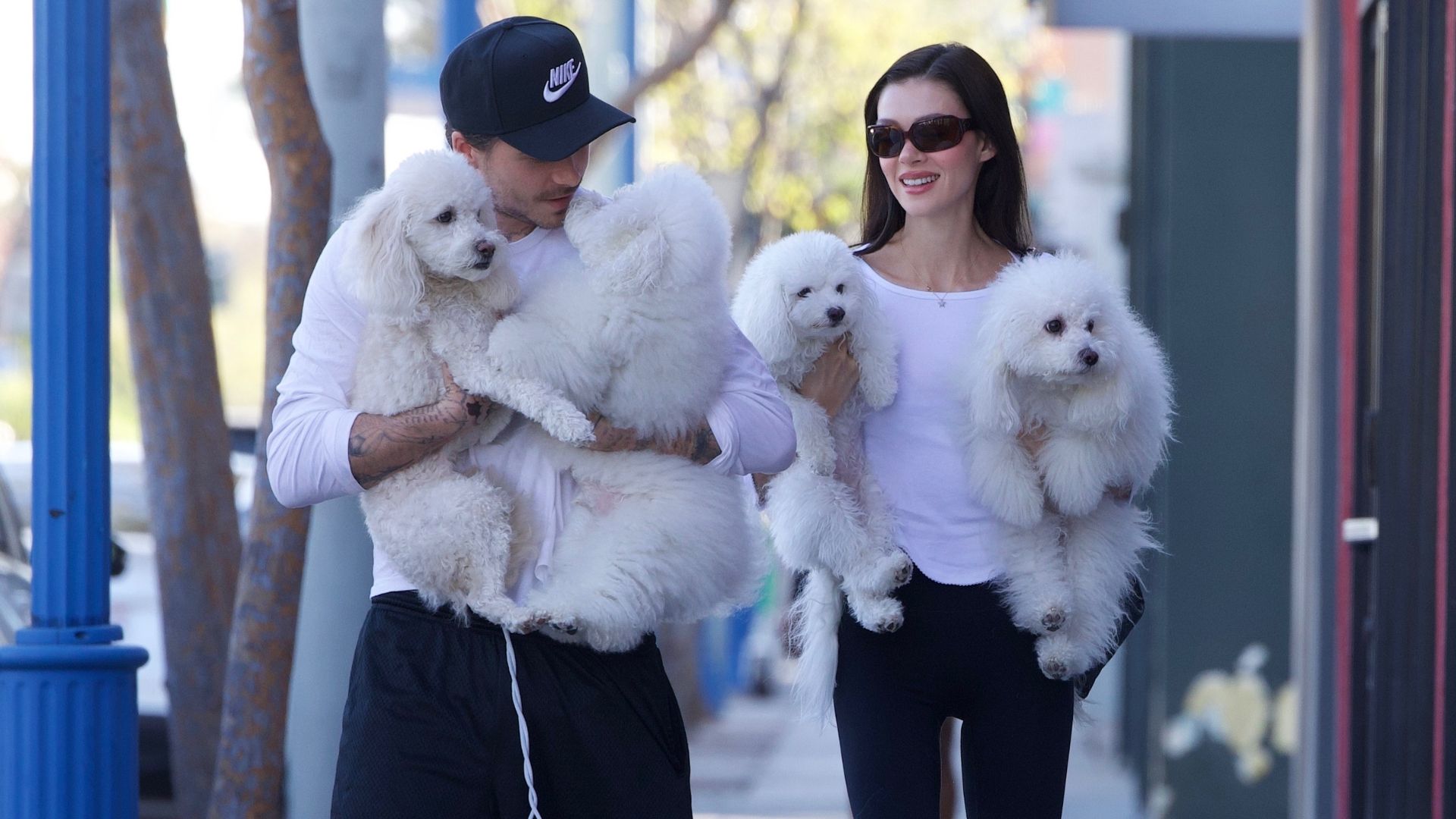 Nicola Peltz Beckham walks alongside Brooklyn Beckham carrying puppies. She wears a white t-shirt with black capri leggings and black rounded-toe heels.