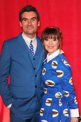 Jeff Hordley with wife Zoe Henry on the red carpet, wearing a blue suit in front of a red cutrain
