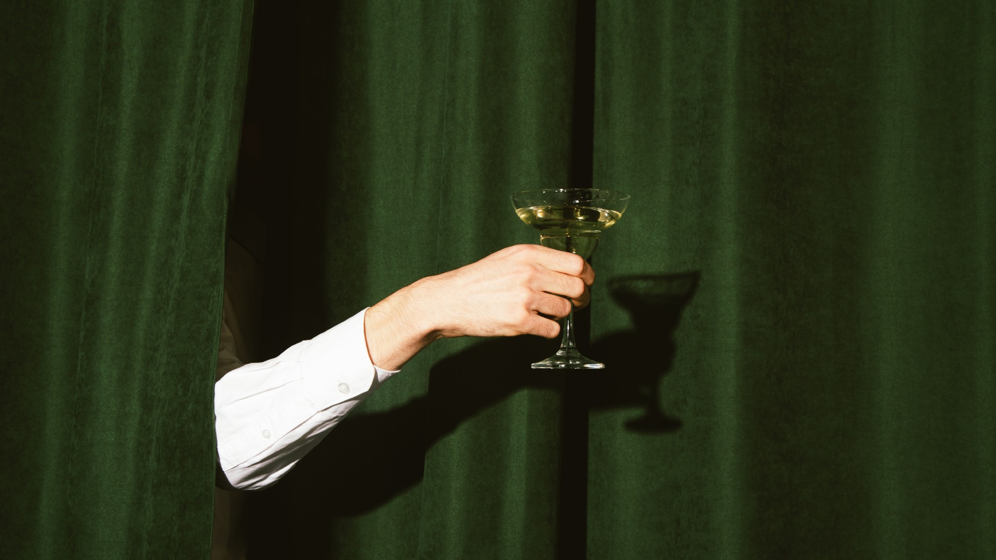 an arm pokes through a green curtain, presenting a martini cocktail in a coupe glass