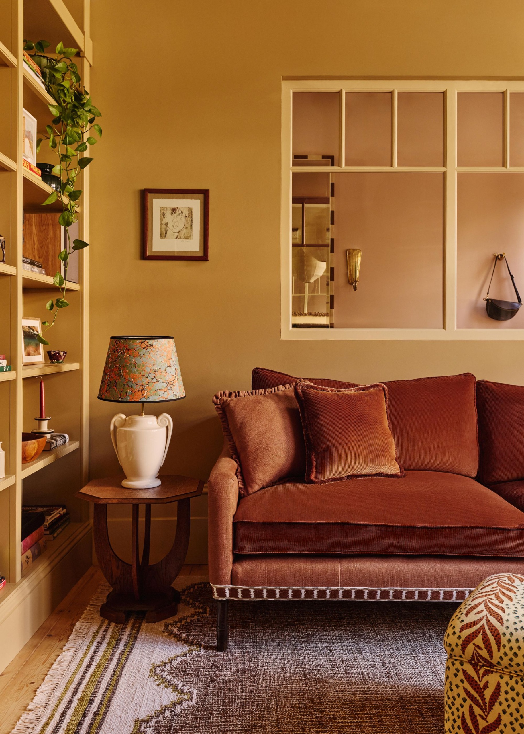 A living room with ochre walls, a window into a pink room, a dark rust velvet sofa, a textured lilac rug, a wall of ochre cabinetry, and a small wooden side table with a lamp.