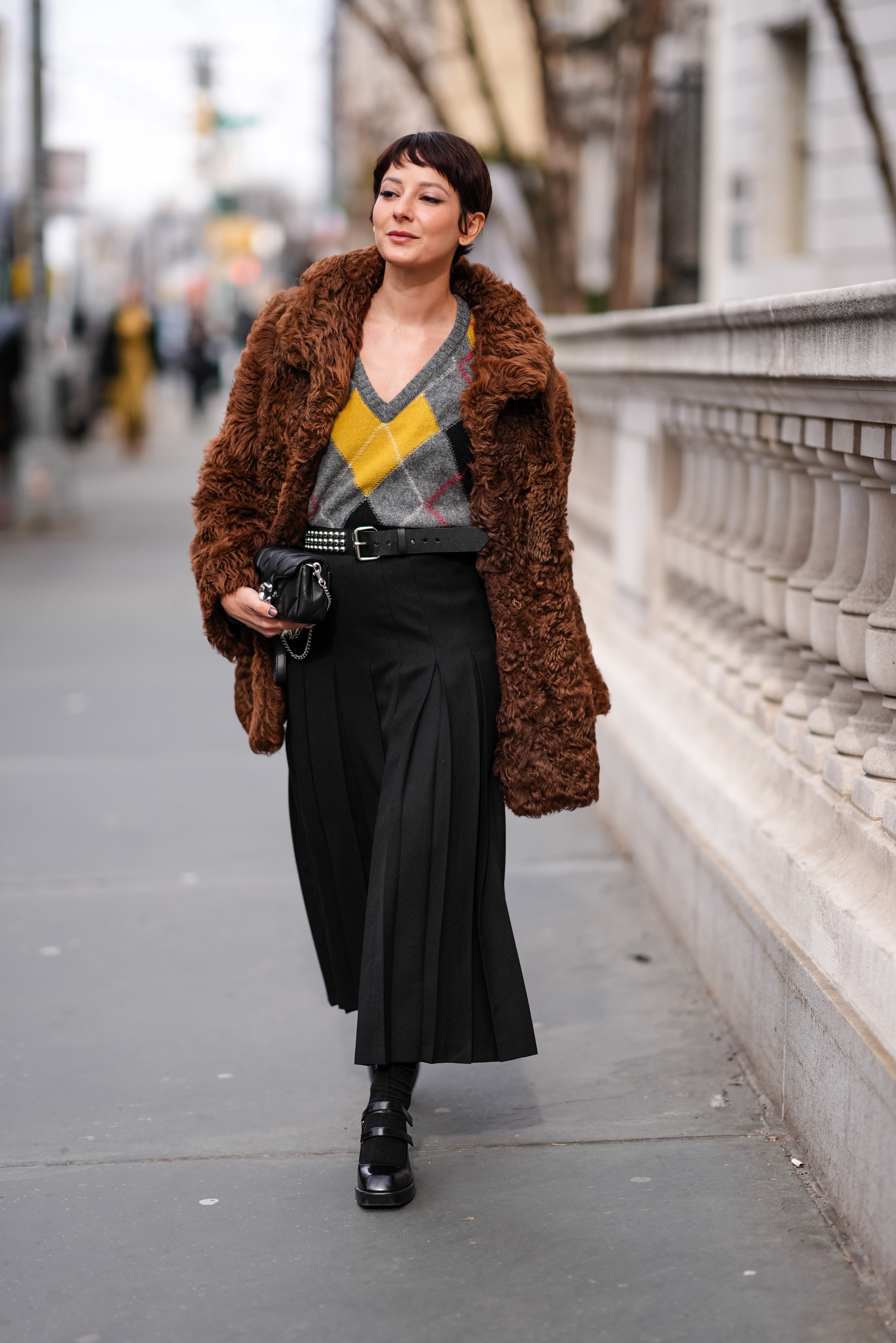 NEW YORK, NEW YORK - FEBRUARY 12: Alyssa Coscarelli wears a brown oversized fluffy jacket, a gray and yellow low-neck v-neck argyle pattern printed pullover, a black midi pleated skirt, black leather high heels shoes, a belt, a black leather Coach bag, outside Coach, during New York Fashion Week, on February 12, 2024 in New York City. (Photo by Edward Berthelot/Getty Images)