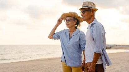 A happy older couple holds hands as they look off into the distance at the beach. 