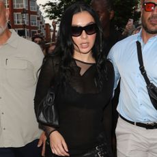 Charli XCX walks down the street in London wearing a black leather miniskirt with black pointed-toe heels and a sheer black top.