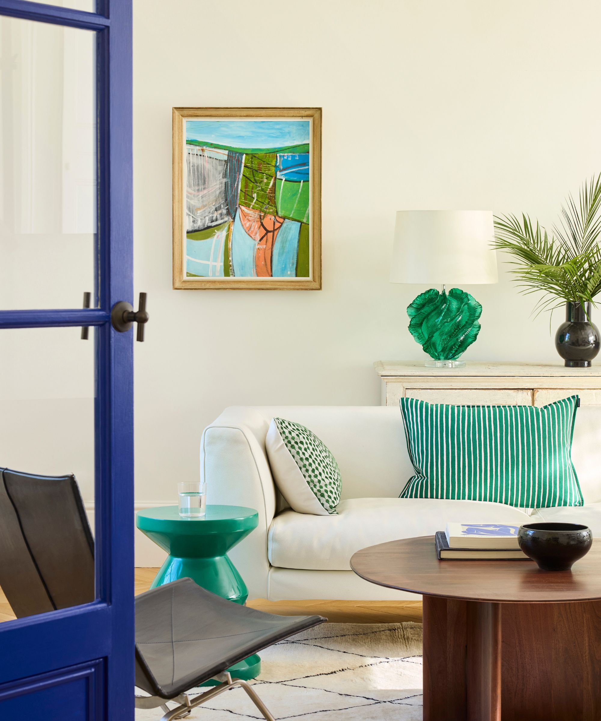 A white living room with a cream sofa, blue and green abstract artwork, green sofa cushions, a round wooden coffee table, and a green side table.