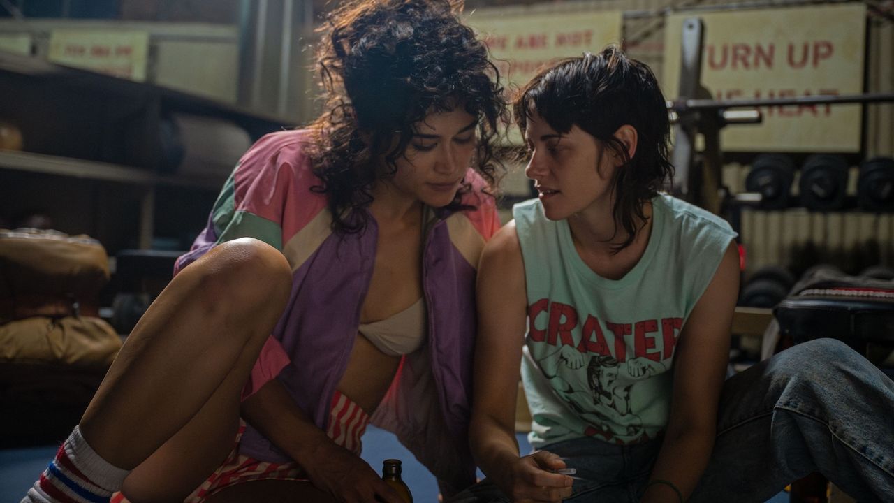 a body builder and a woman in a muscle t-shirt sit on the floor of a gym in a still from love lies bleeding