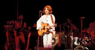 George Harrison wears white and plays acoustic guitar onstage in 1974