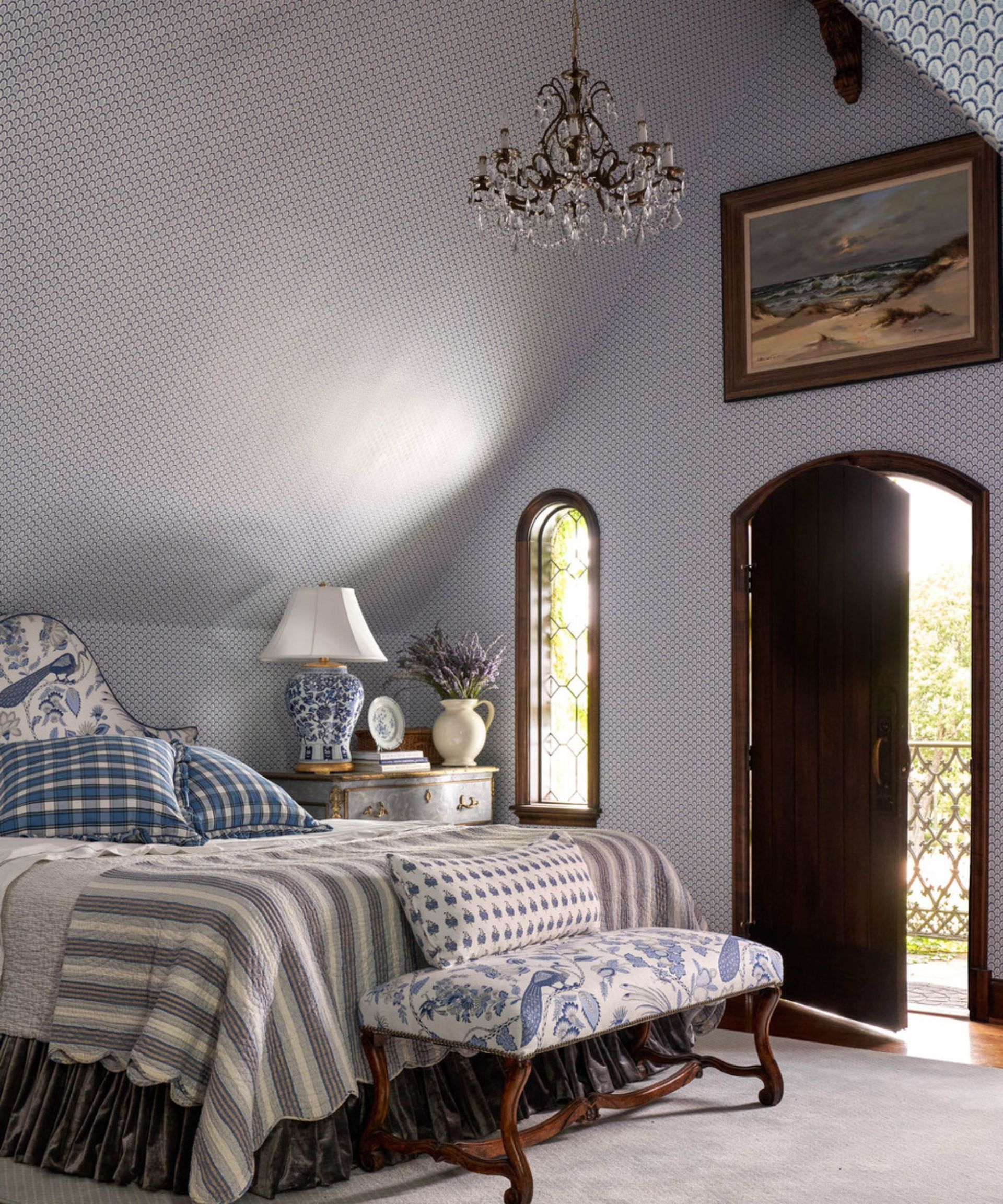 Bedroom with high ceiling, arched door with balcony, chandelier, blue and white bedding and upholstered footstool
