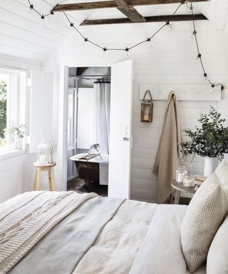 A cream bedroom with ensuite bathroom in background, small bathtub and festoon black bedroom fairy lighting