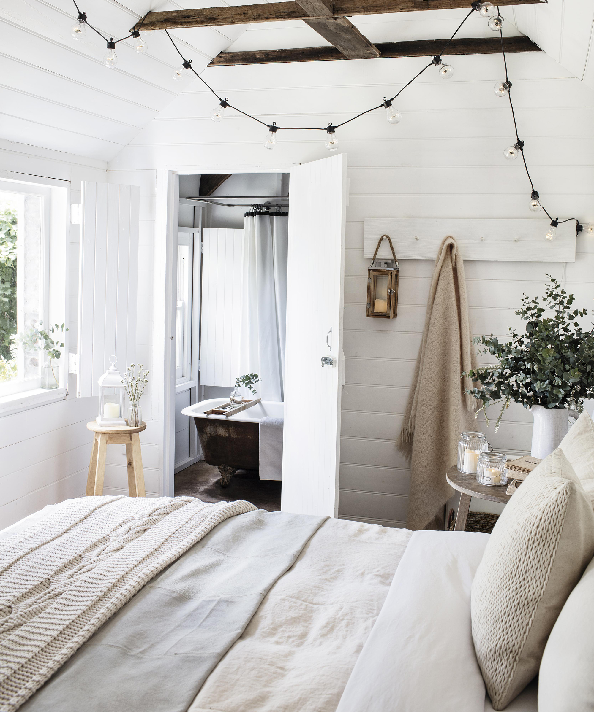 A cream bedroom with ensuite bathroom in background, small bathtub and festoon black bedroom fairy lighting