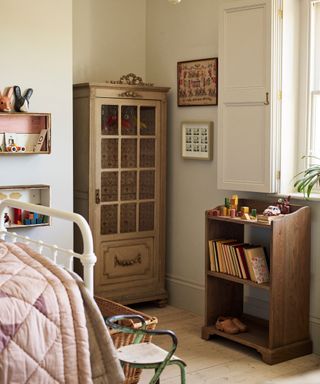 A child's bedroom with a small wooden closet with glass doors, and a wooden bookshelf with books and toys stacked on top. The bottom of a white metal bed visible on the left hand side.