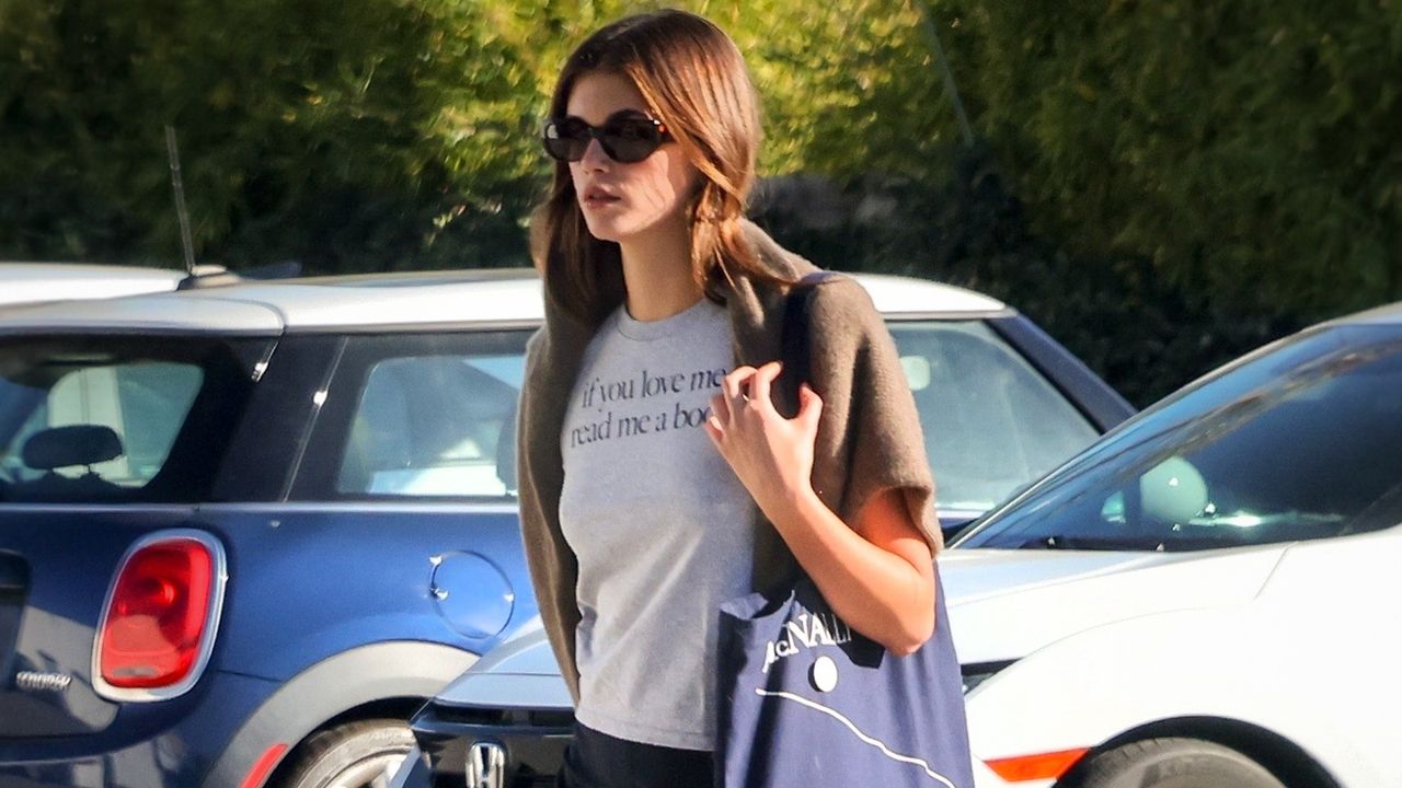 Kaia Gerber wearing a grey tee-shirt, sunglasses, and a blue tote bag