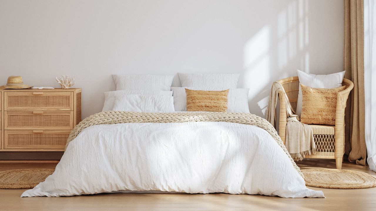 Here is a bedroom with white walls, a bed with white sheets and beige throw pillows and bed runners, a wooden nightstand and chair, and a wooden floor