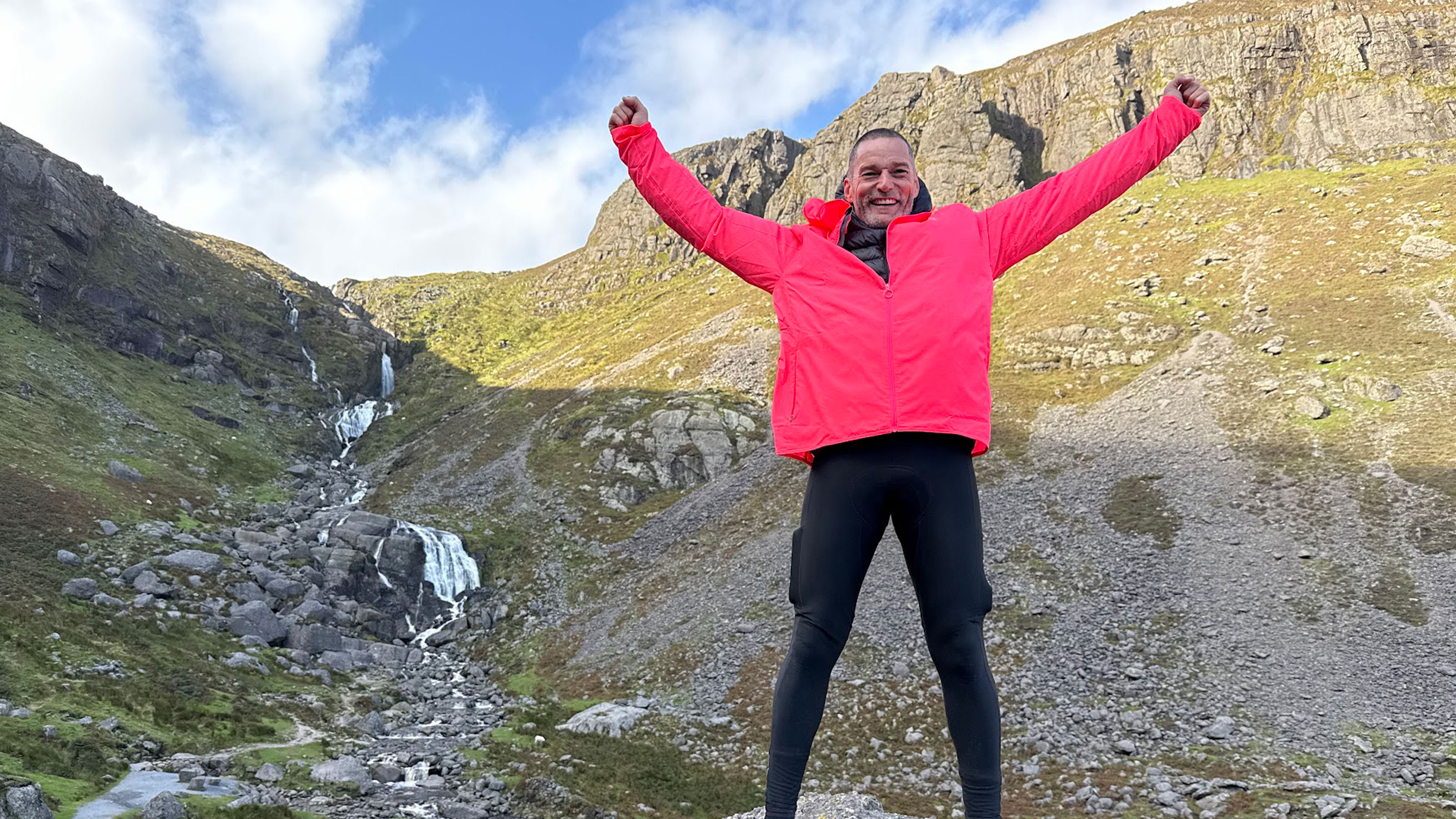 Fred Sirieix in a pink jacket and spandex leggings infront of a rocky landscape with a stream.