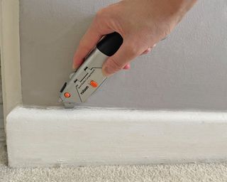 a hand cutting through the caulk between an old skirting board and a wall with a utility knife