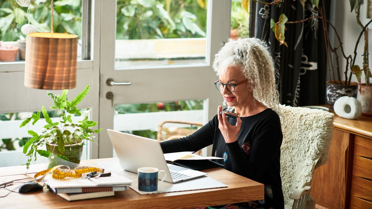 A woman sitting in a light-filled home office talks on a smartphone while sitting at a laptop.