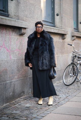 what to wear in 30-degree temperatures, shown in a photo of a woman standing outside wearing a black fur coat over a black ribbed top with a black wool skirt, white boots, and a crescent bag