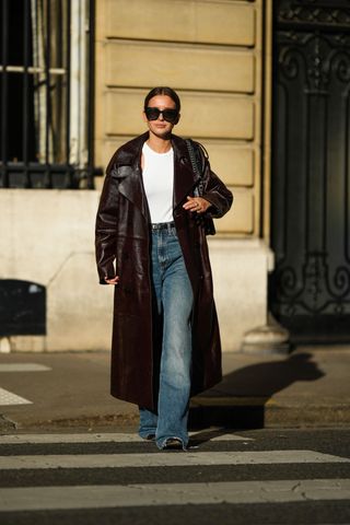 Diane Batoukina wears black sunglasses, a white ribbed cropped tank-top, a dark brown shiny varnished leather long coat, a black grained leather small Kelly belt from Hermes, blue faded denim large ripped pants, black shiny leather shoes , during a street style fashion photo session, on October 26, 2022 in Paris, France.