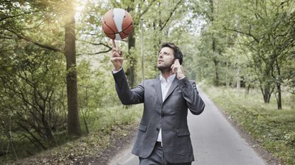 A businessman talks on the phone while spinning a basketball on his finger on a woodsy walking path.