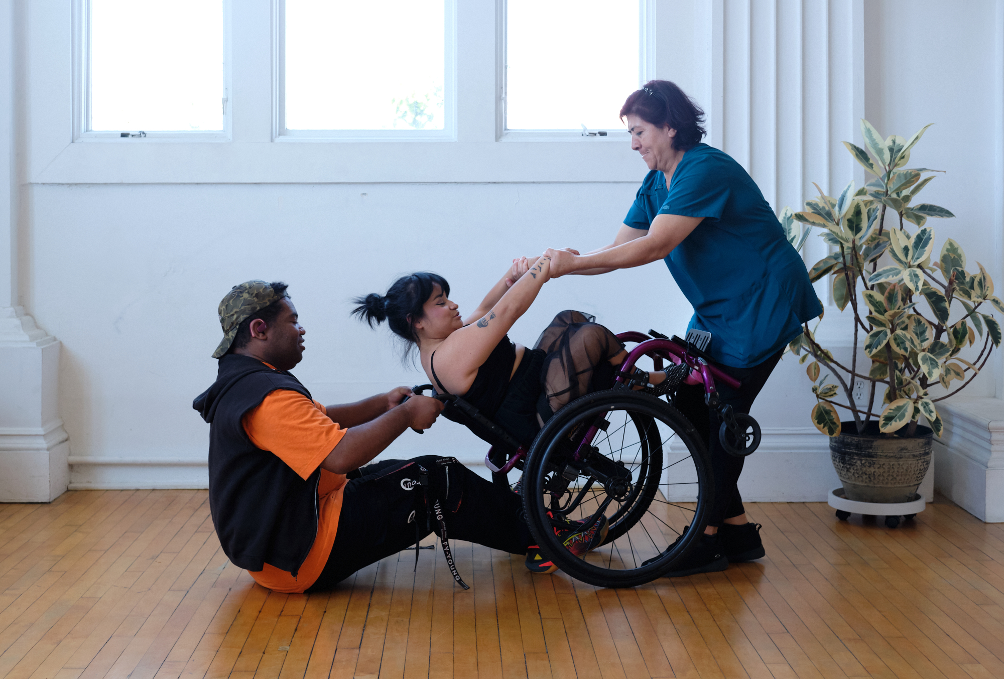 Three people engage in a collaborative movement exercise in a bright room, with two individuals assisting a person seated in a wheelchair.