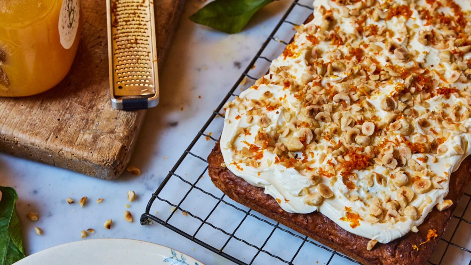 Parsnip cake with white frosting and nuts on top on a wire rack with a zester and pot of honey to the side