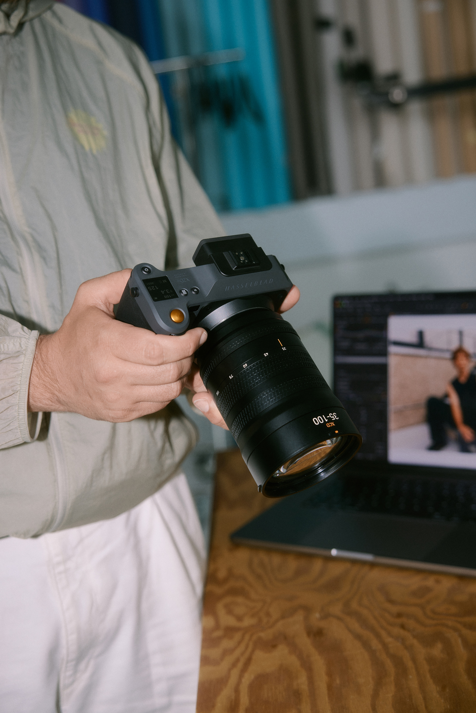 Person holding a Hasselblad camera with a 35-100mm lens, standing near a laptop showing a blurred photo editing screen