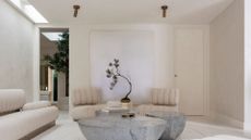 A white living room with white ribbed sofa seaters and a couch, alongside a trio of stone coffee tables, a stack of books, and a potted mini tree