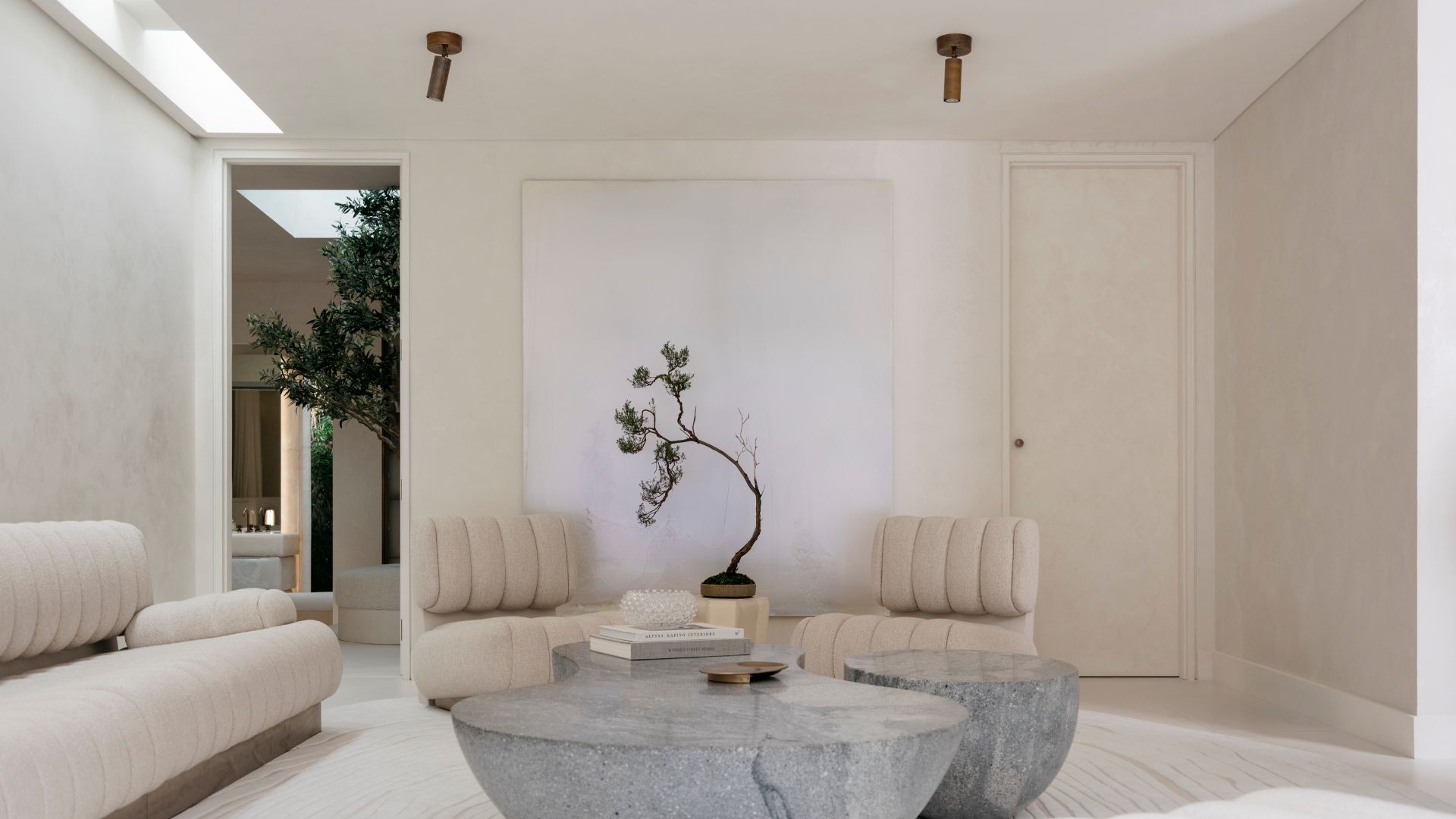 A white living room with white ribbed sofa seaters and a couch, alongside a trio of stone coffee tables, a stack of books, and a potted mini tree