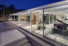 Southridge glass house exterior, stone steps to the side/front of the building, white interior, furniture and plants inside, black piano to the right, spot light ceiling, patio area with white chairs, blue night sky, surrounding landscape of trees and rocky area