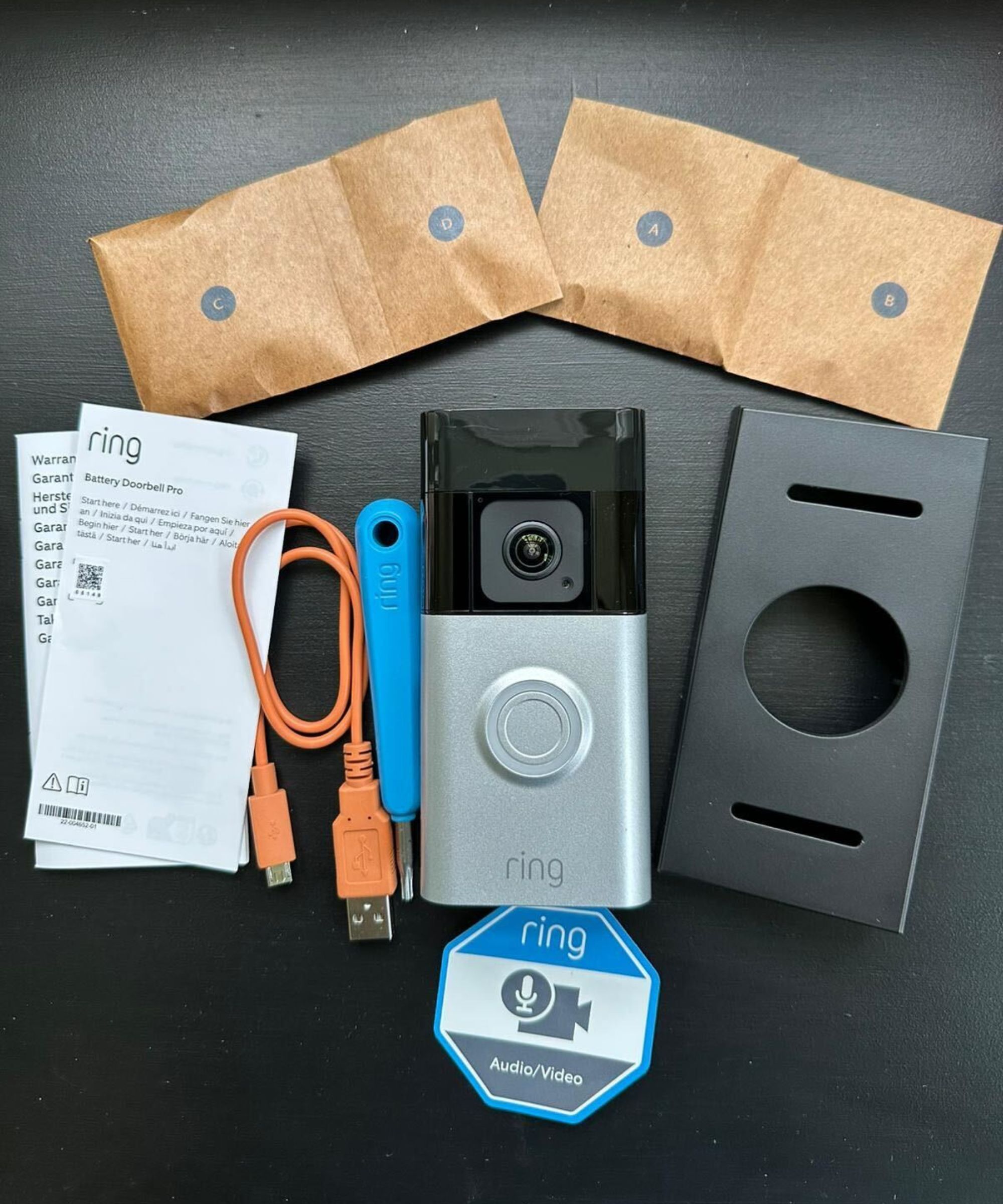 A silver and black Ring Battery Doorbell Pro unboxed on a black table, with a black mount, blue screwdriver, orange charging cable, two instruction manuals, four small brown paper envelopes, and a hexagonal white, blue and gray ring audio/video sticker.
