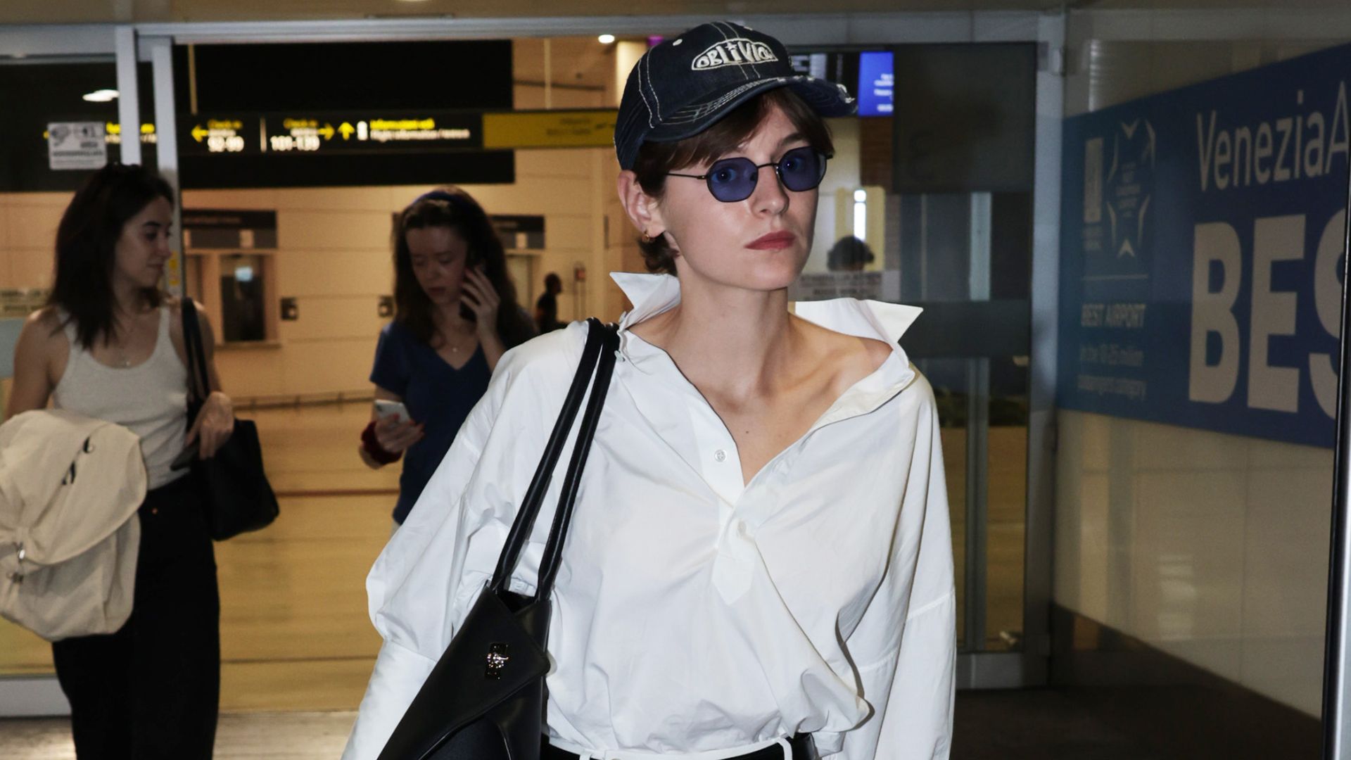 Emma Corrin walks through the airport wearing an all-white outfit with a black bag, black belt and black Derby shoes.