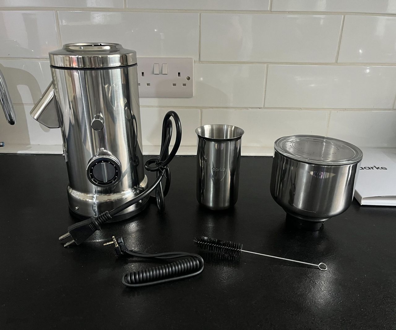 The parts of the Aarke coffee ginder, including the mechanical base, stainless steel grounds cup, shopper with glass lid, cleaning brush, and connector cable, on a black counter.
