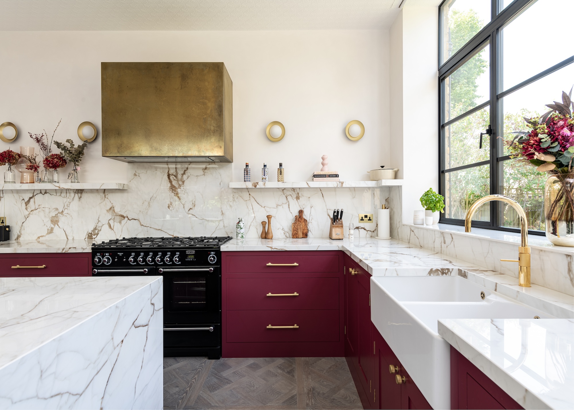 Deep red lower kitchen cabinets with veined marble worktops and open shelving. There is also a large island and brass colored extraction fan