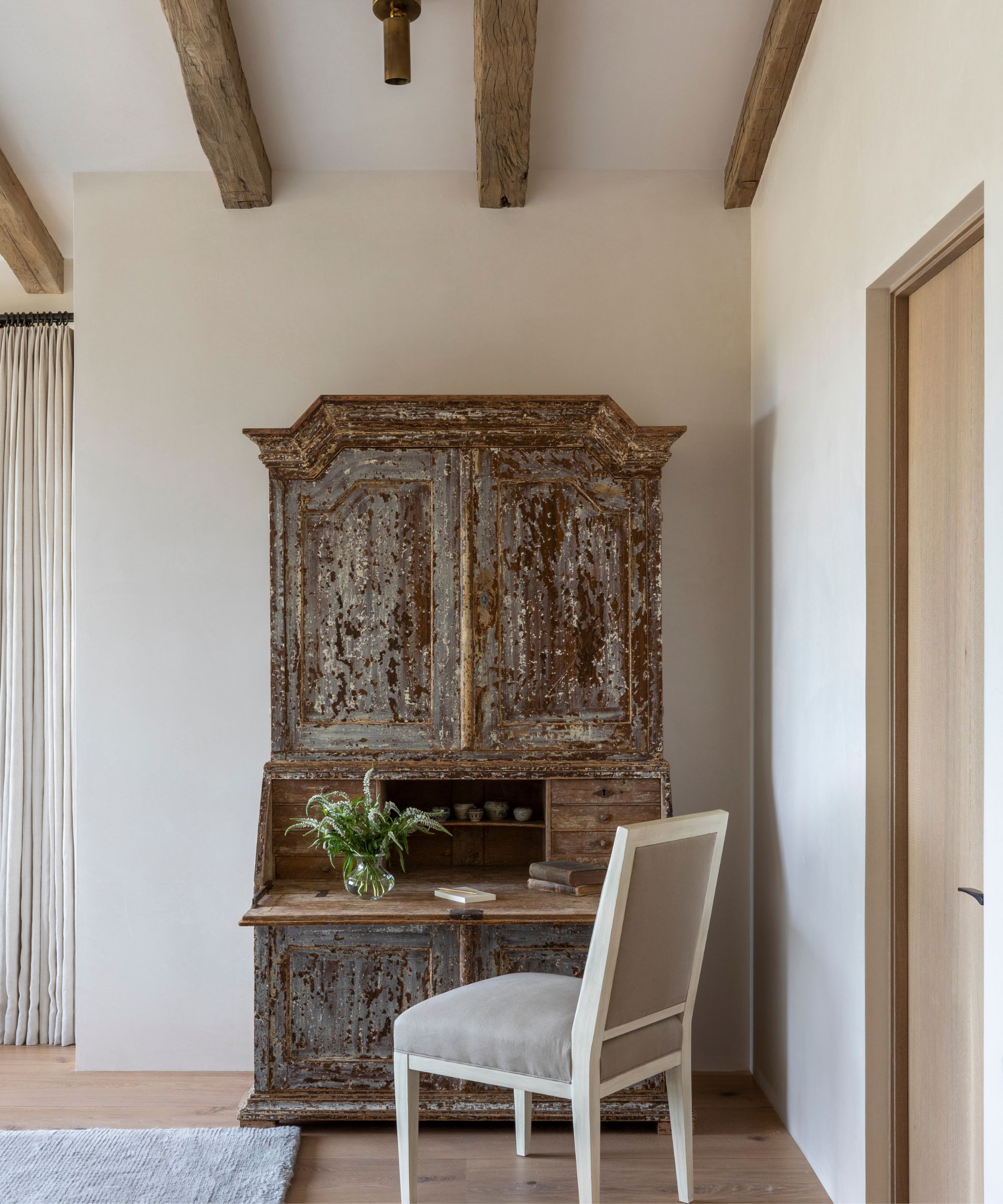 Home office with antique distressed writing desk opened out to reveal storage and a vase of foliage, a white upholstered dining chair, beamed ceiling, cream walls, and a wooden floor.