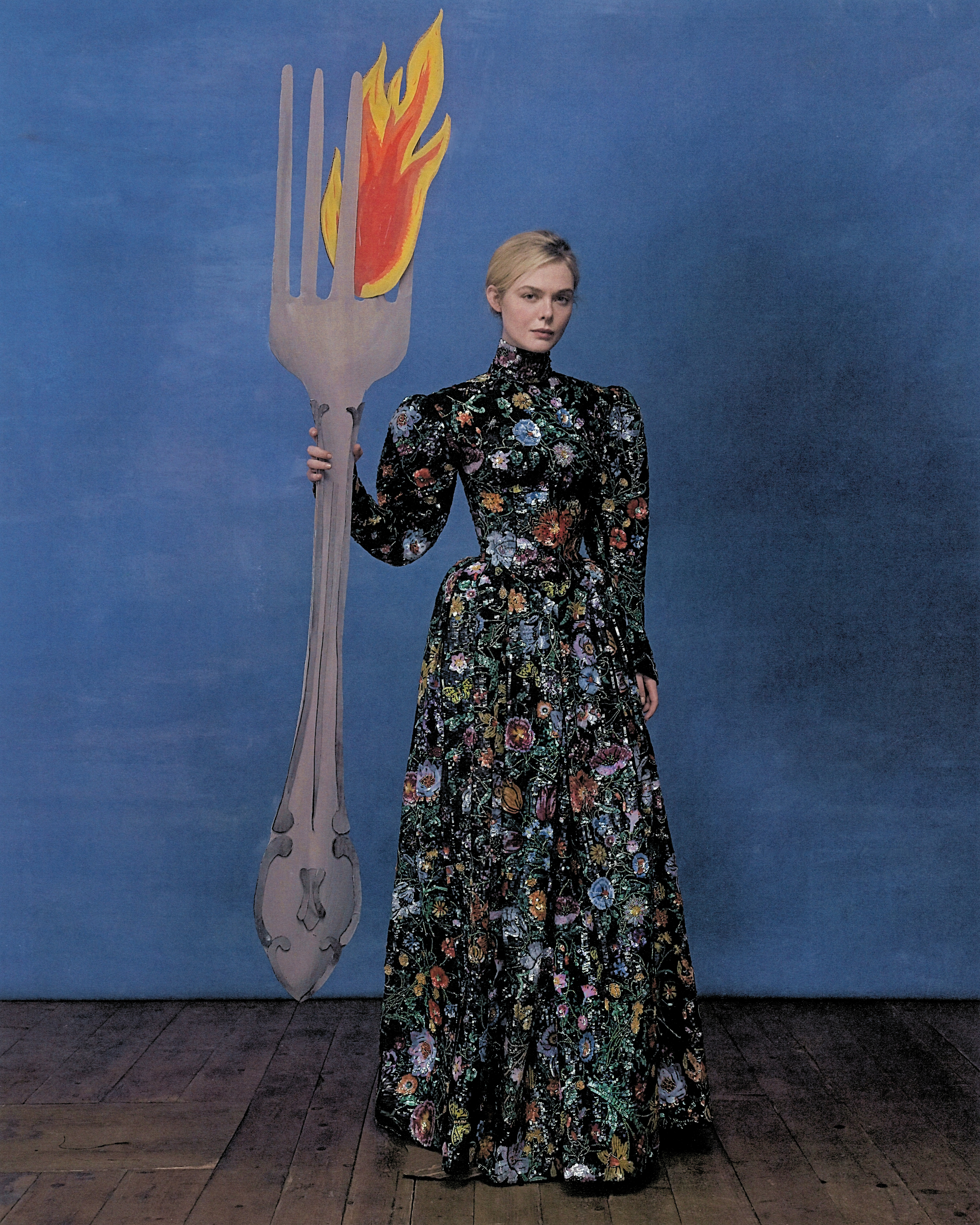 Elle Fanning shot by Szilveszter Mak&amp;oacute;. Fanning is wearing a long sleeved Gucci gown with embroidered florals and is seen holding a large life-size cardboard cutout of a fork.