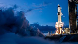 a black and white spacex rocket test-fires its first-stage engines on the launch pad beneath a dark-blue late-evening or early-morning sky