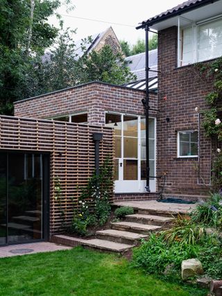 Daytime exterior view of a brick and timber house extension, green lawn, stone steps, flowers and shrubs, black frame patio window, black drainpipe, white framed glazed door, window, tall trees in the backdrop, pale sky