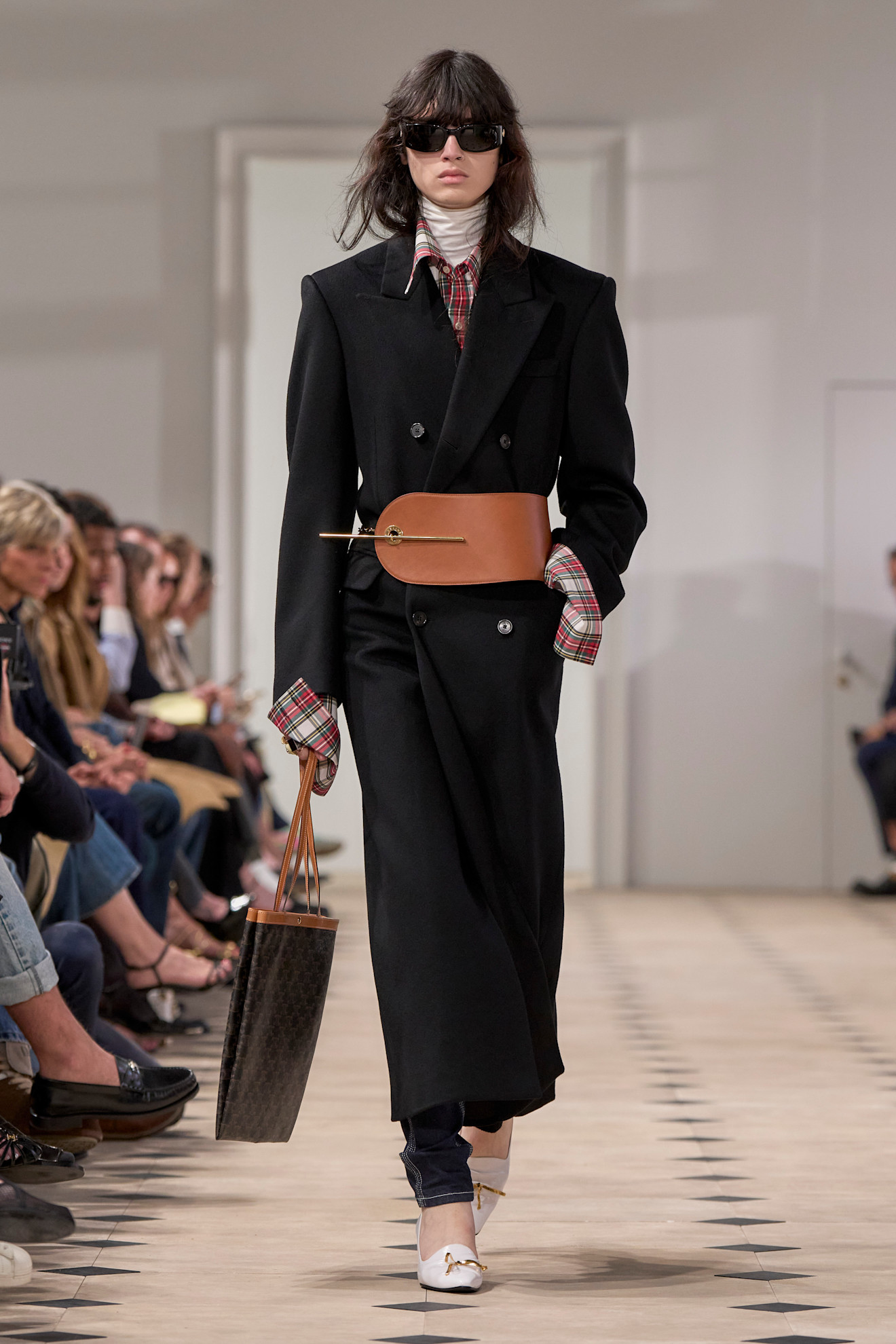Layering staples for spring 2026 is shown in a photo from Celine's runway show, with a model walking while wearing a long black coat with a large tan corset belt layered on top. Underneath the model is wearing a red flannel shirt on top of a white turtleneck styled with dark wash skinny jeans, white pointed pumps, and a brown tote bag