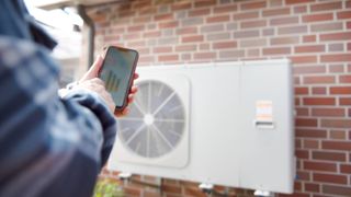 person using phone app to look at graph with air source heat pump on wall of house in background