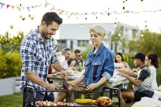 A man and a woman are cooking food on a BBQ in the garden with friends.