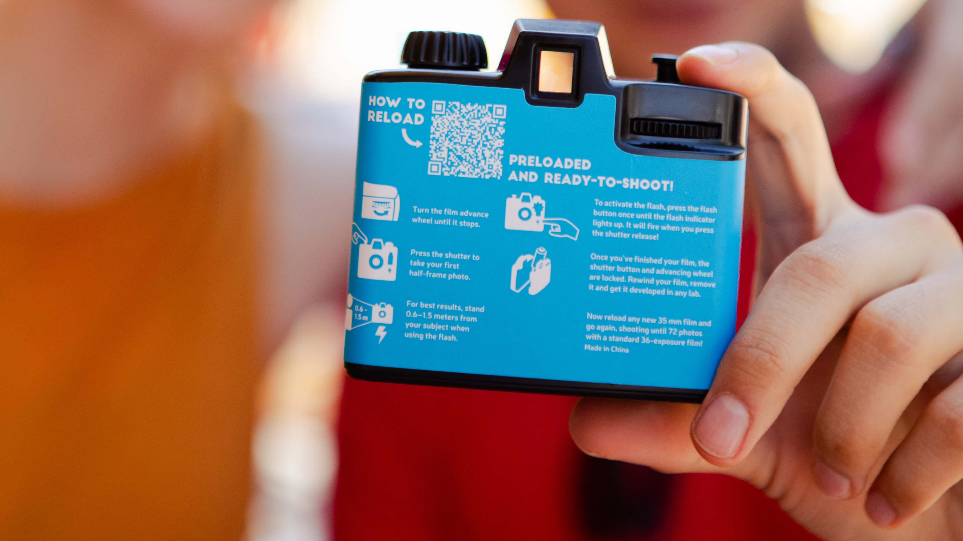 Photograph of a hand holding the Lomography Half-frame Simple Use Reloadable Film Camera, showing the rear of the camera with written instructions, diagrams and a QR code