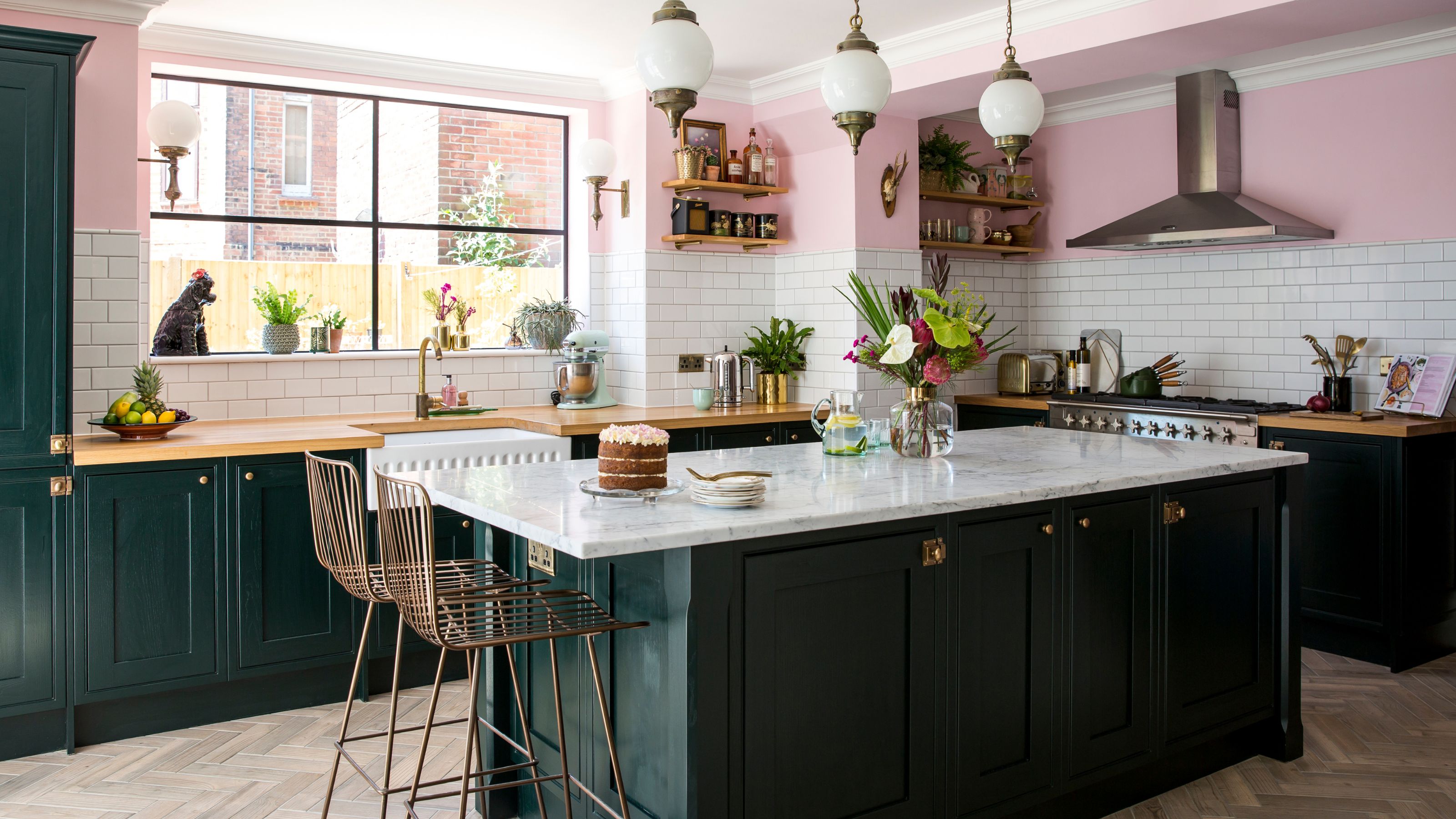 Basement kitchen with dark green units, pink walls and white metro tiles, with an island in the centre of the space