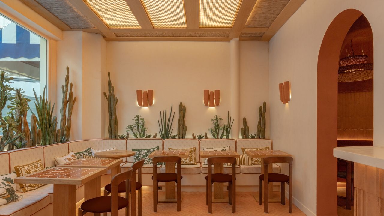 A modernly furnished, adobe architecture-inspired restaurant features soft, warm lighting, wooden and rattan furniture, and decorated fabric banquettes.