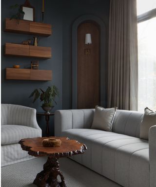 dark blue painted bedroom with a large window and a seating area created below with a small grey sofa, a striped loose linen armchair and a vintage coffee table