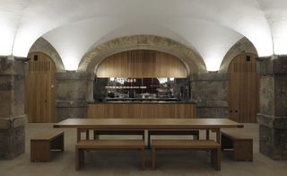 Christ Church Crypt, Spitalfields, by Dow Jones Architects, stone wall and pillars, large wooden table and benches, arched white ceiling with up lights, wooden counter top with mirror wall section behind, rounded wooden doors, neutral stone floor