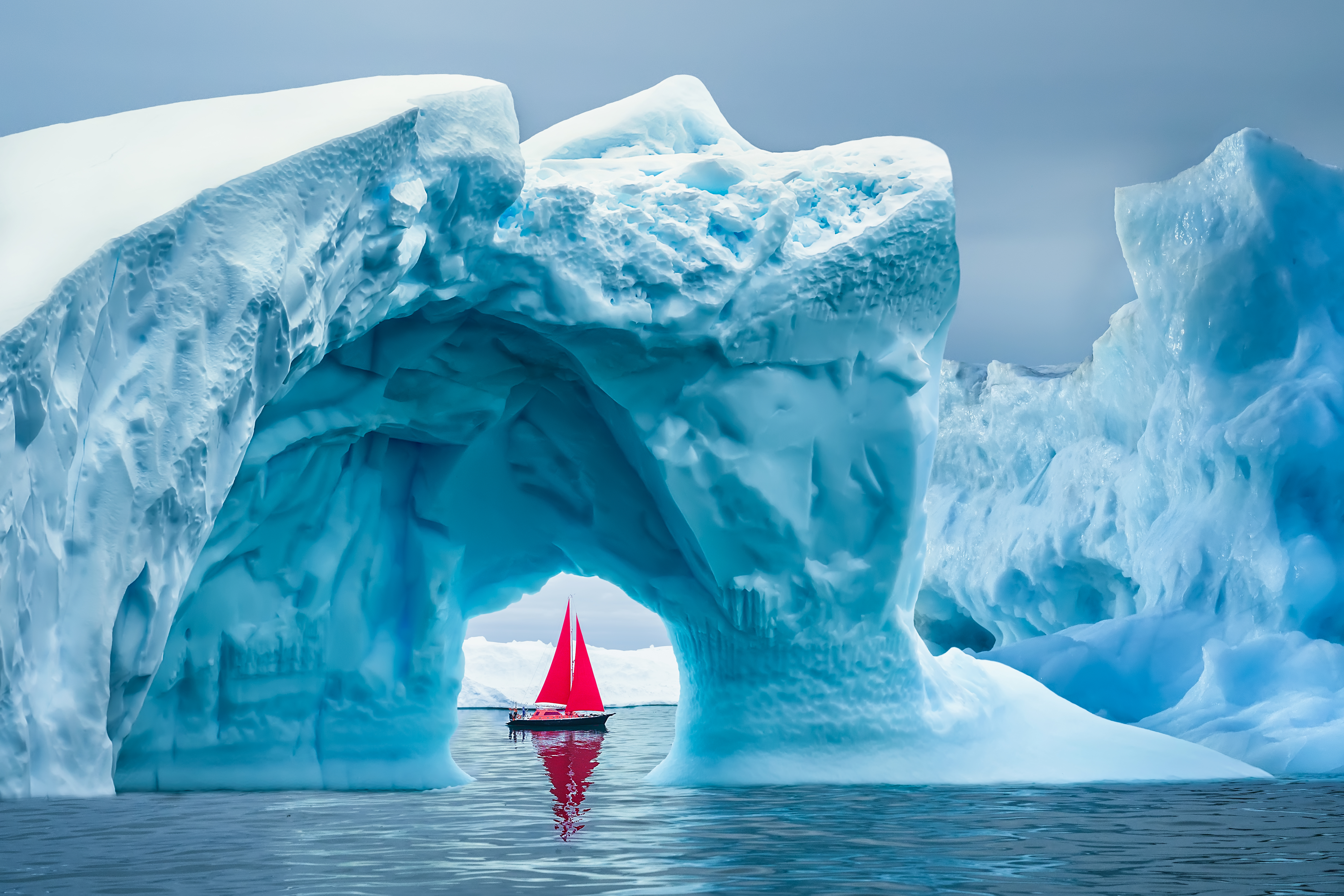 Boat with red sails in the distance, framed by large iceberg archway with blue sky above