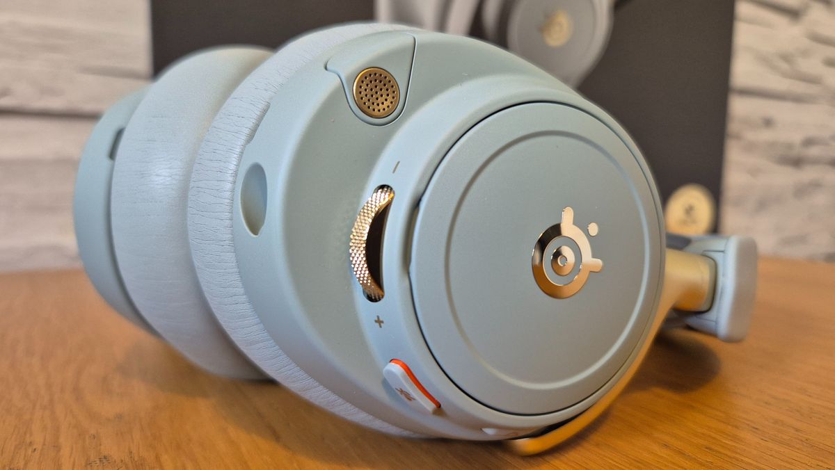 The SteelSeries Arctis Nova Elite headset in front of a white brick background and on a wooden surface