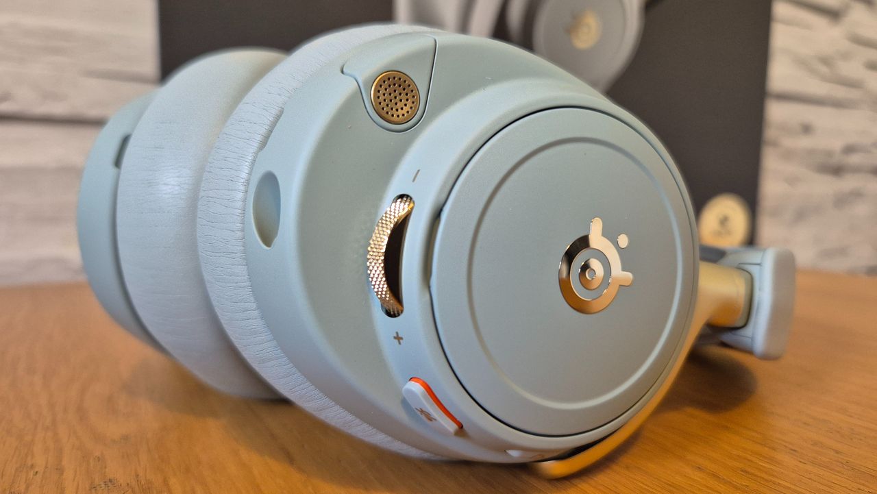 The SteelSeries Arctis Nova Elite headset in front of a white brick background and on a wooden surface