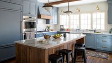 A transitional kitchen with blue-gray cabinets, a wooden island with white countertops, black bar stools, and wood ceiling beams. Brass wall sconces by the window and a butler's sink. 
